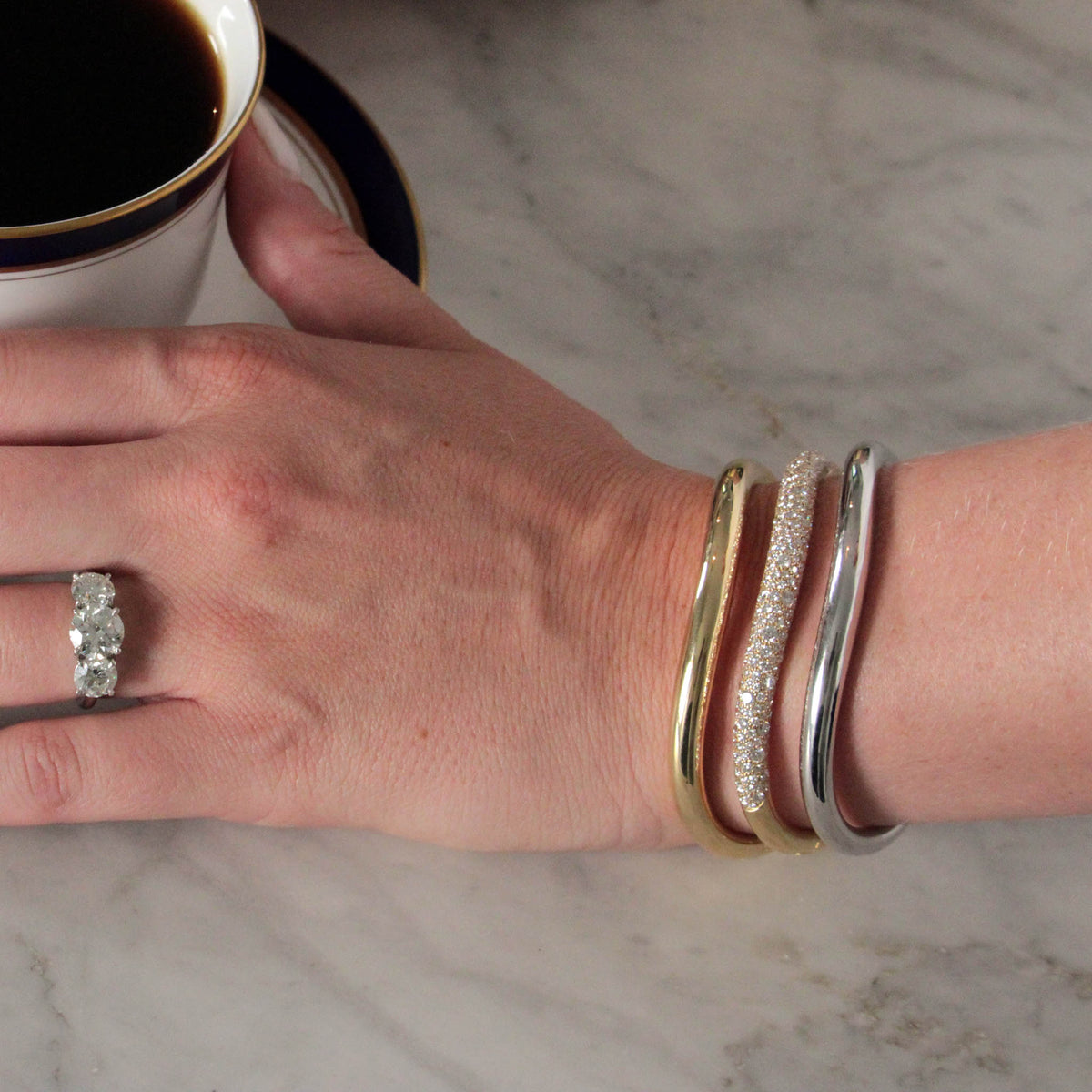 hand with coffee cup wearing three-stone diamond engagement ring and triple stack of paul morelli bangles