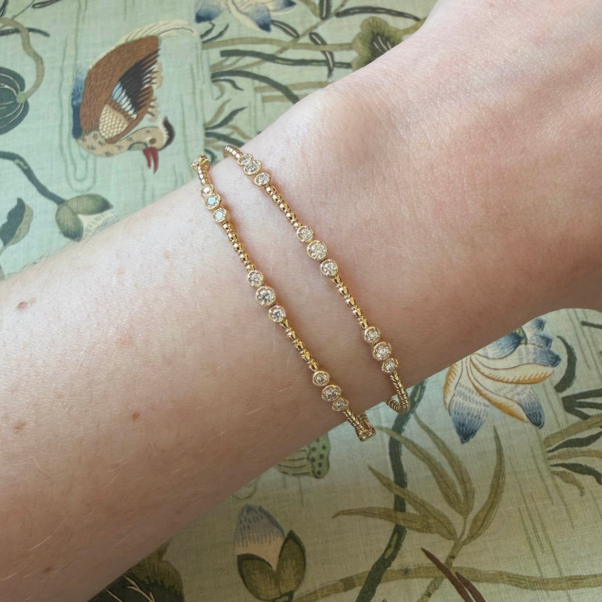 Diamond Triple Bezel Milgrain 5-Station and 3-Station 14K Yellow Gold Flex Bangles on a wrist over a patterned background.