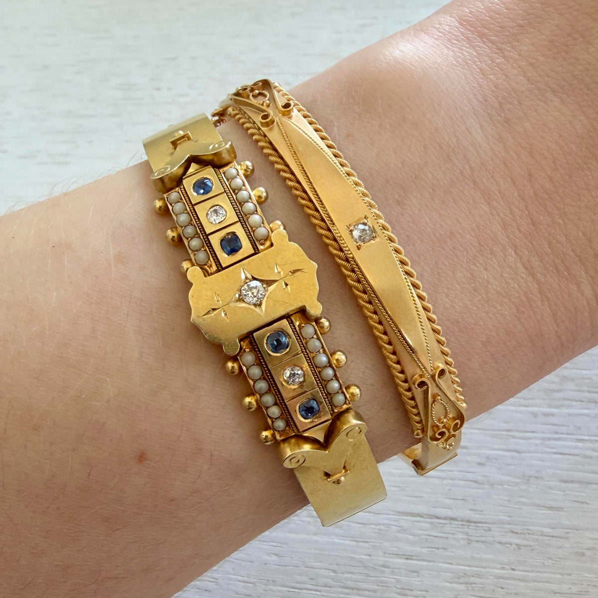 Victorian diamond, sapphire, and pearl gold bangle worn on a wrist beside another gold diamond bangle.