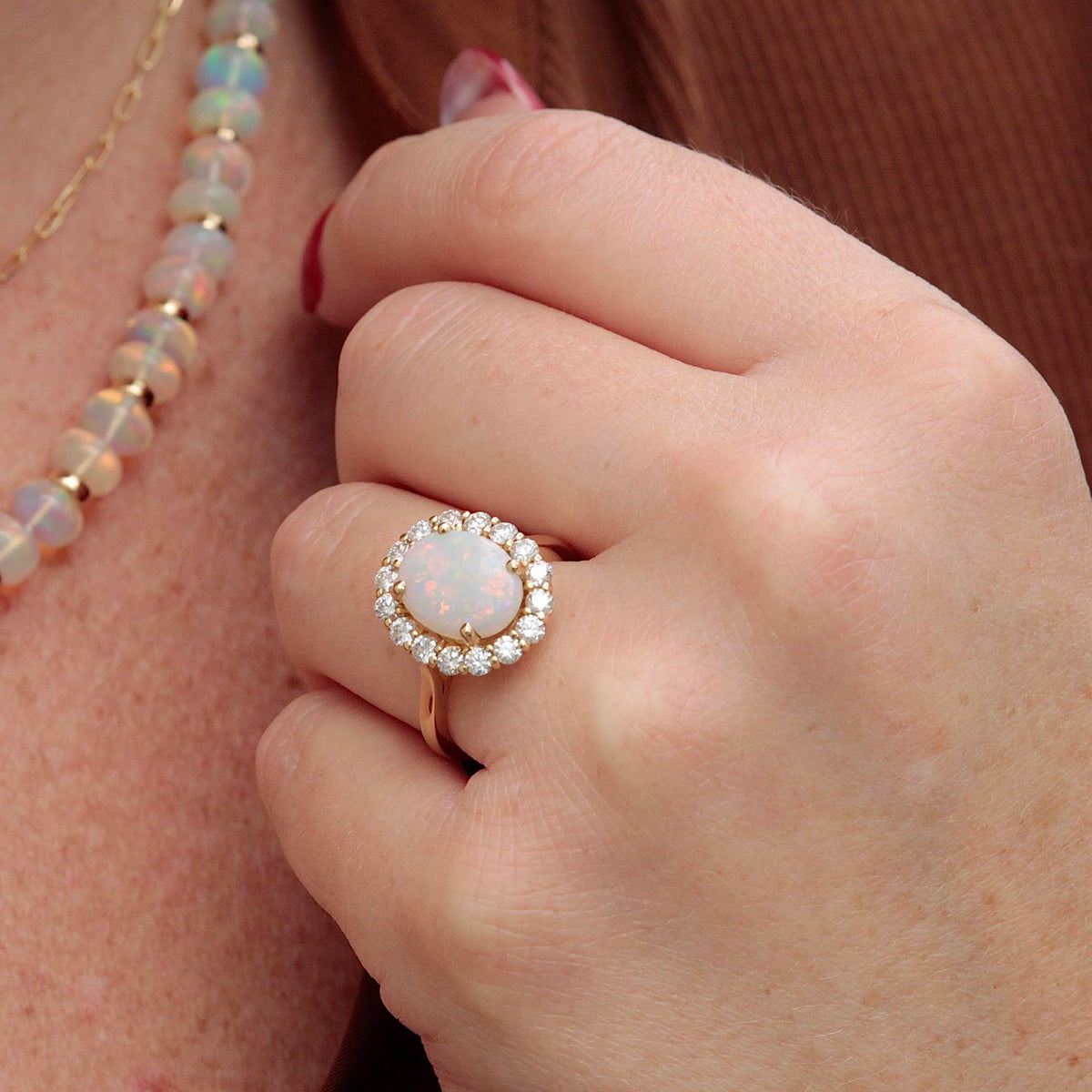 Oval opal and diamond cluster ring in 14K yellow gold worn on a hand beside an opal bead necklace.