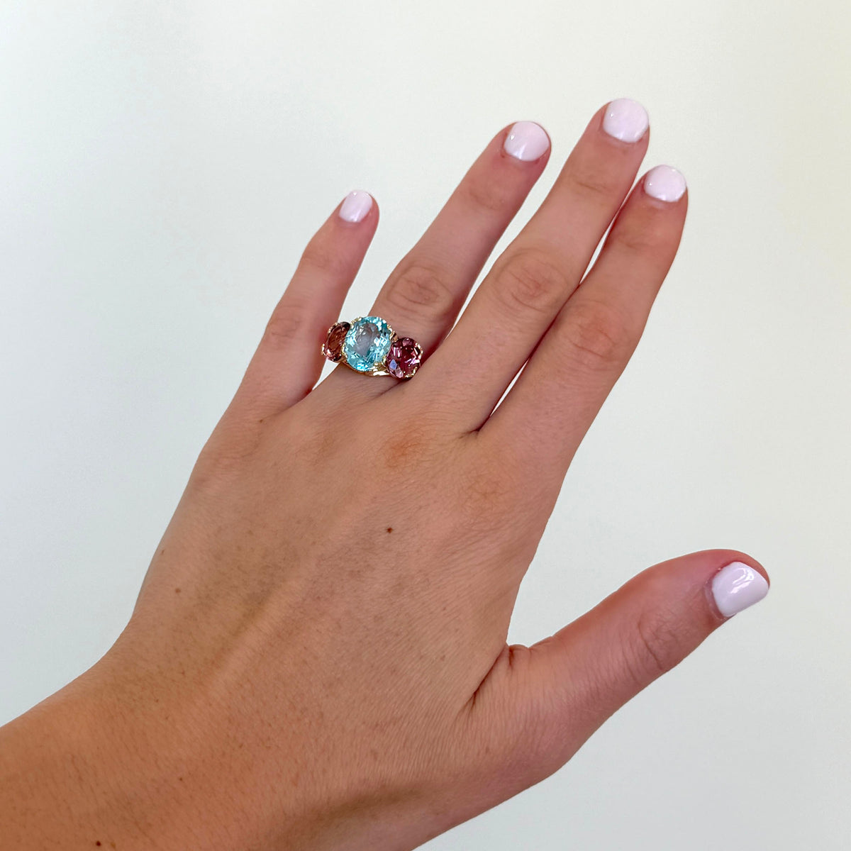 A close-up of a hand wearing a three-stone ring with a central light blue oval gemstone flanked by two small round pinkish-red gemstones, set in a gold-toned band, against a plain off-white background.