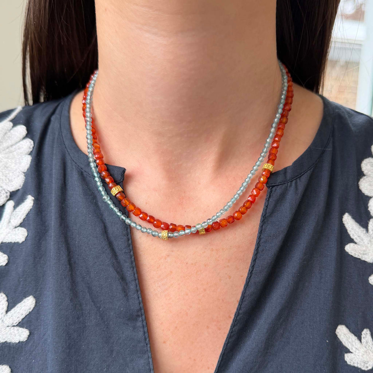 Model wearing a Tippy Stern faceted blue zircon necklace with a yellow gold rondelle, layered with a Tippy Stern orange carnelian necklace with yellow gold rondelles.