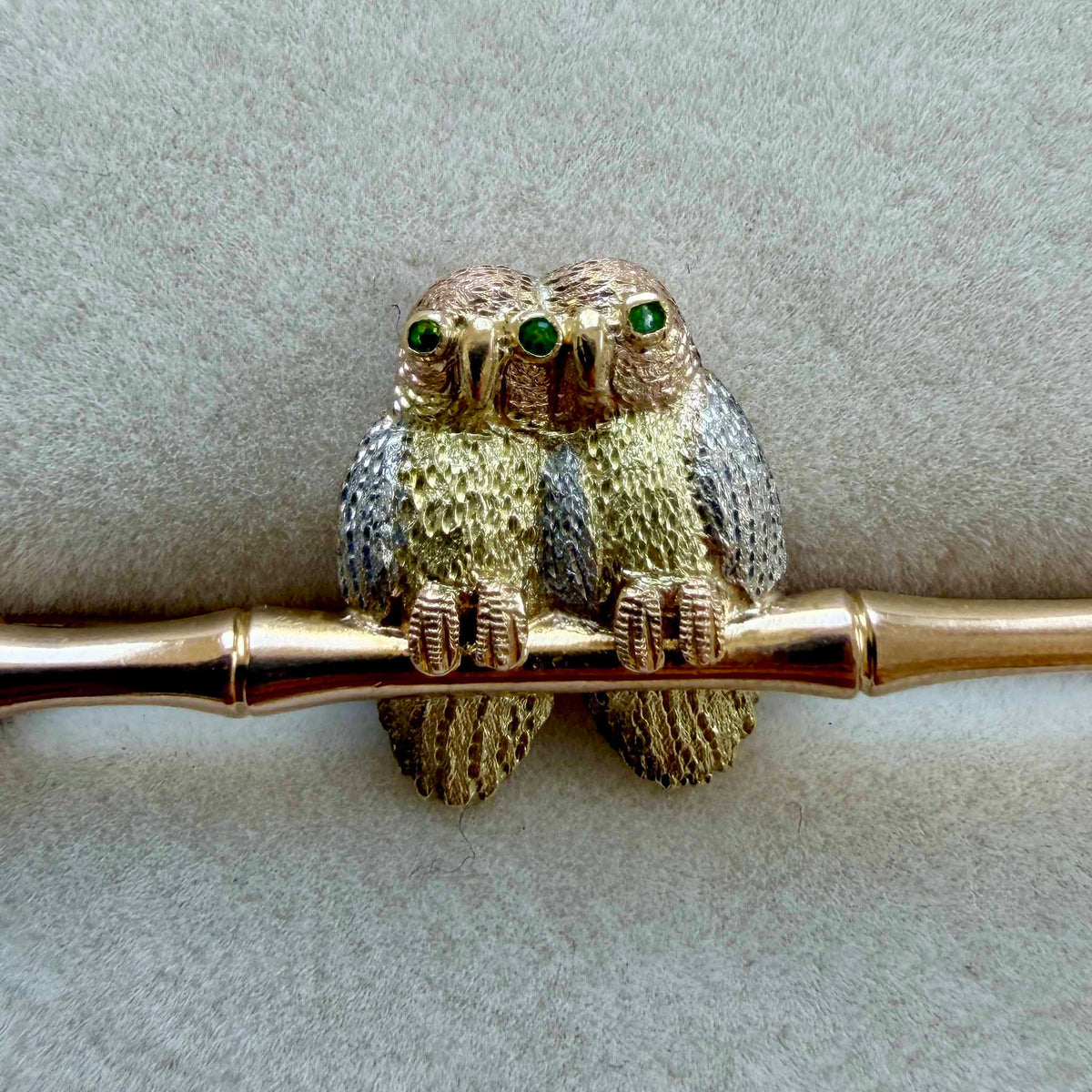 Close-up of two owls on a vintage bamboo pin, showing green eyes, gold-toned bodies, and silver-toned wings.