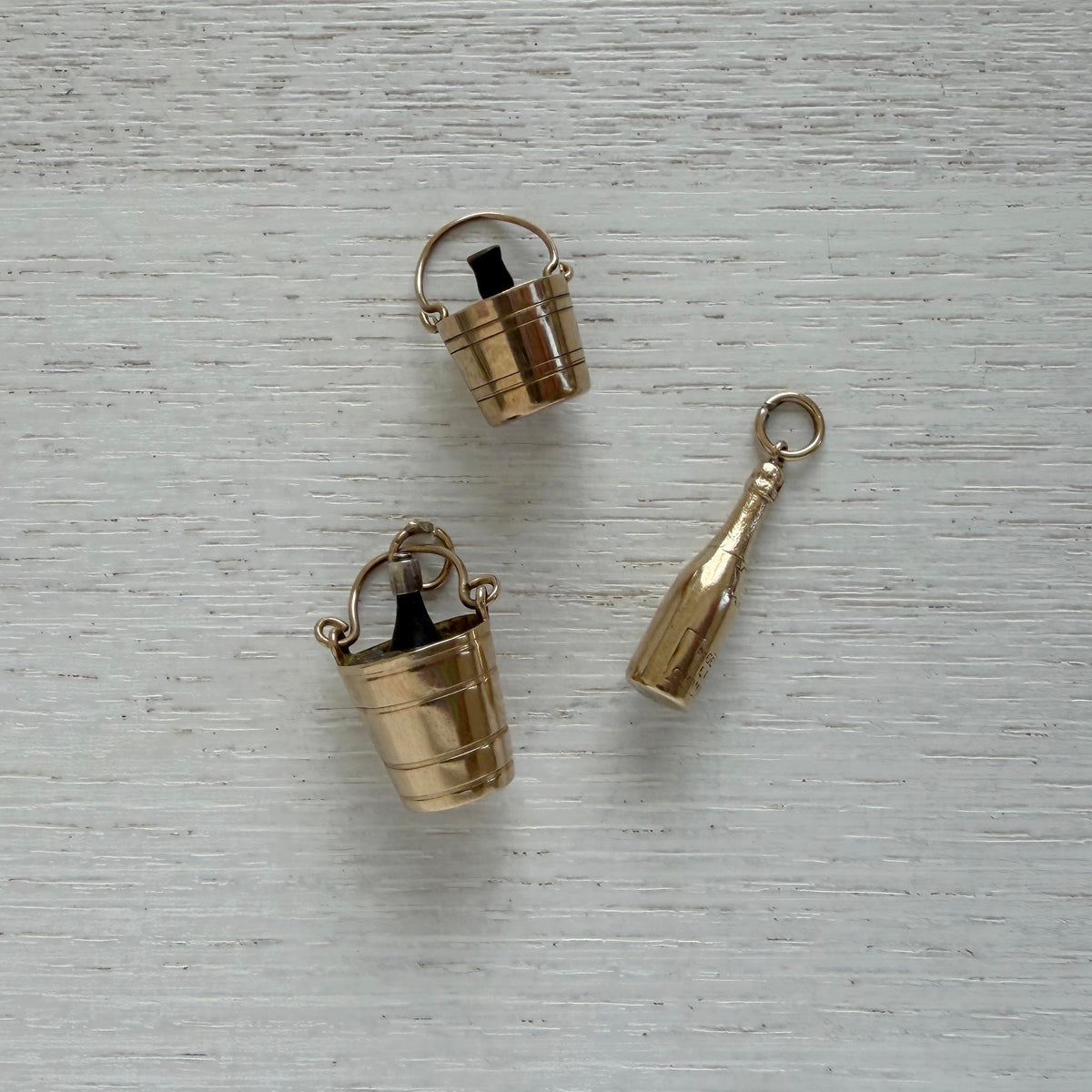 Vintage gold champagne bucket charm displayed beside another bucket charm and a champagne bottle charm on white wood.