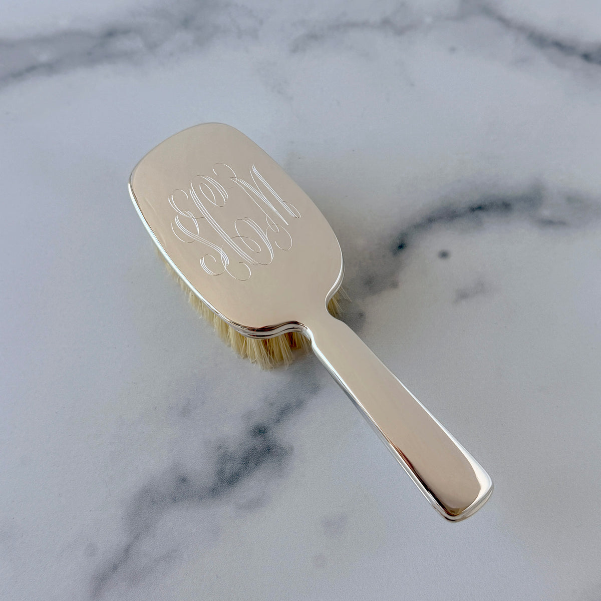 A polished gold hairbrush with a monogrammed face, resting diagonally on a white and gray marble surface.