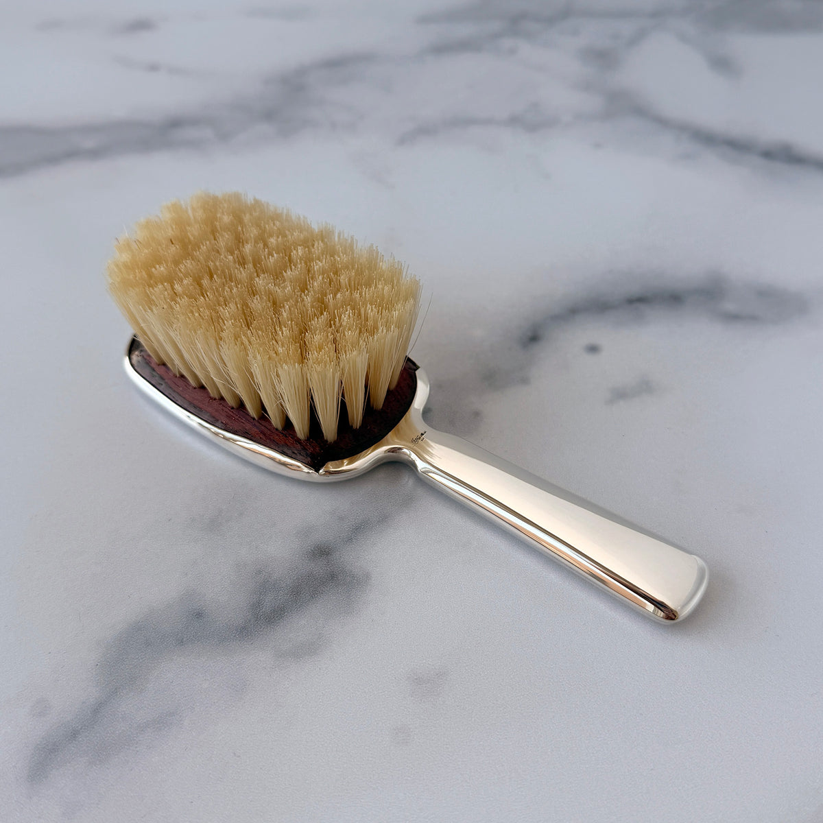 A small grooming brush with a polished silver handle and head, beige natural bristles, and a dark reddish-brown underside, resting on a white marble surface.