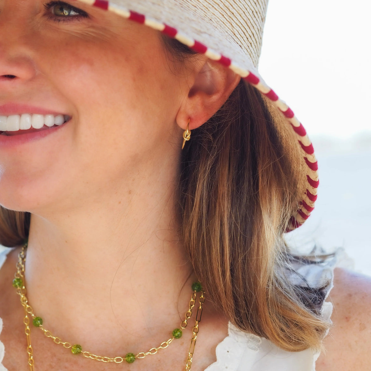 A close-up lifestyle shot of a woman wearing a straw sun hat with a red and white striped brim, layered gold necklaces, and a small gold hoop earring.