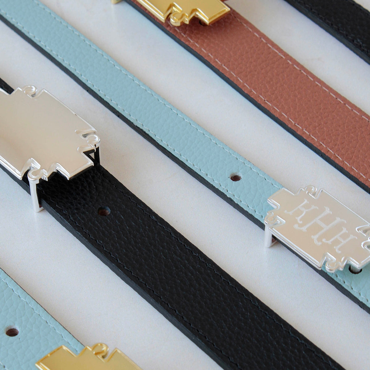 Flat lay of leather belt straps in light blue, black, and brown with silver and gold plaque buckles, one engraved with a monogram.
