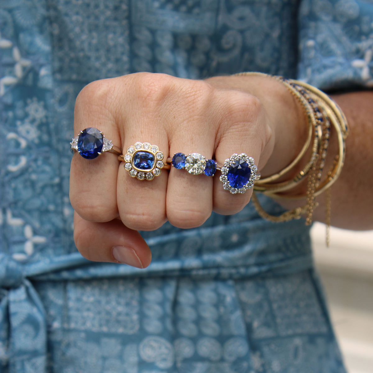 A close-up of a hand wearing four rings with blue sapphires and clear stones, along with stacked gold bangles, against a blue patterned dress background.