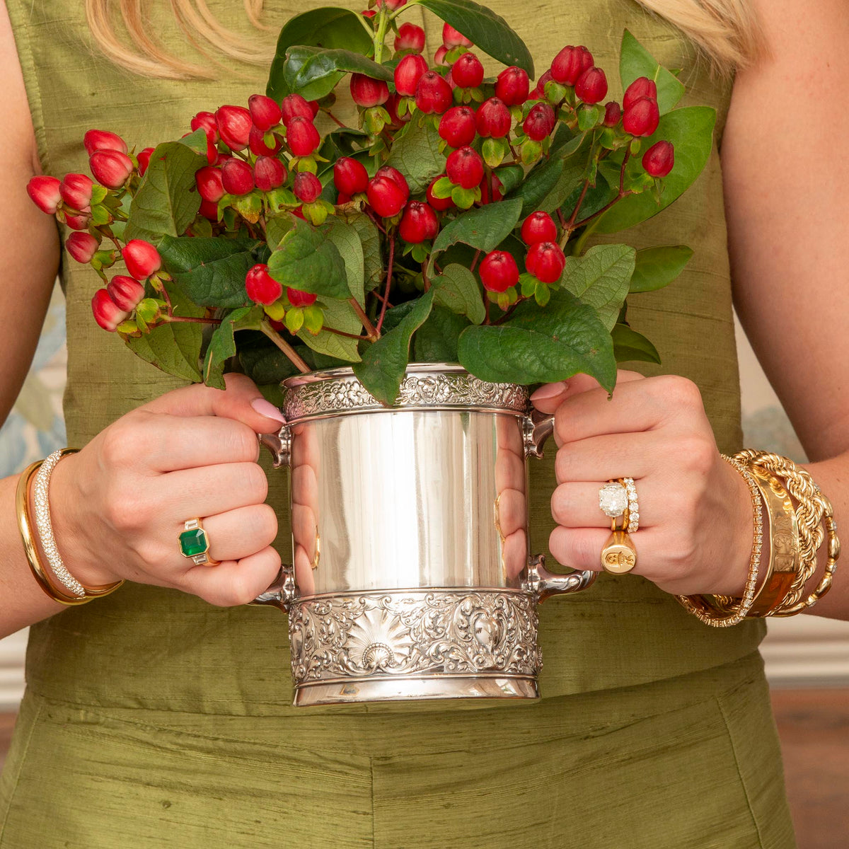 A close-up lifestyle shot of a woman in an olive green dress holding a silver-toned metal mug filled with red berries and green leaves.