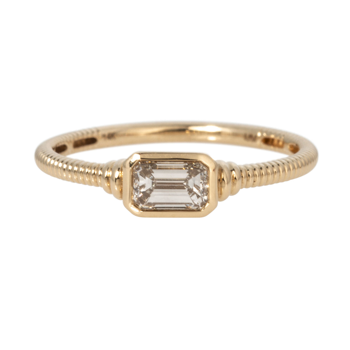 East-west emerald-cut diamond solitaire ring with a bezel setting and ribbed 14K yellow gold band.