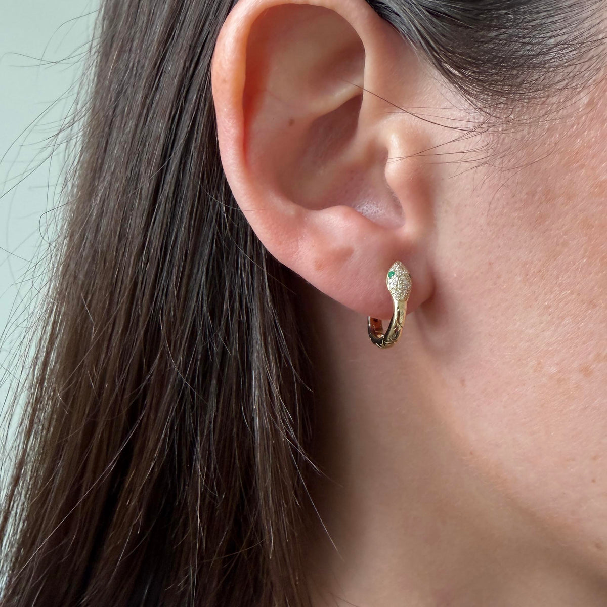 Model wearing a 14K yellow gold diamond and emerald snake huggie earring, showing its fit around the earlobe.