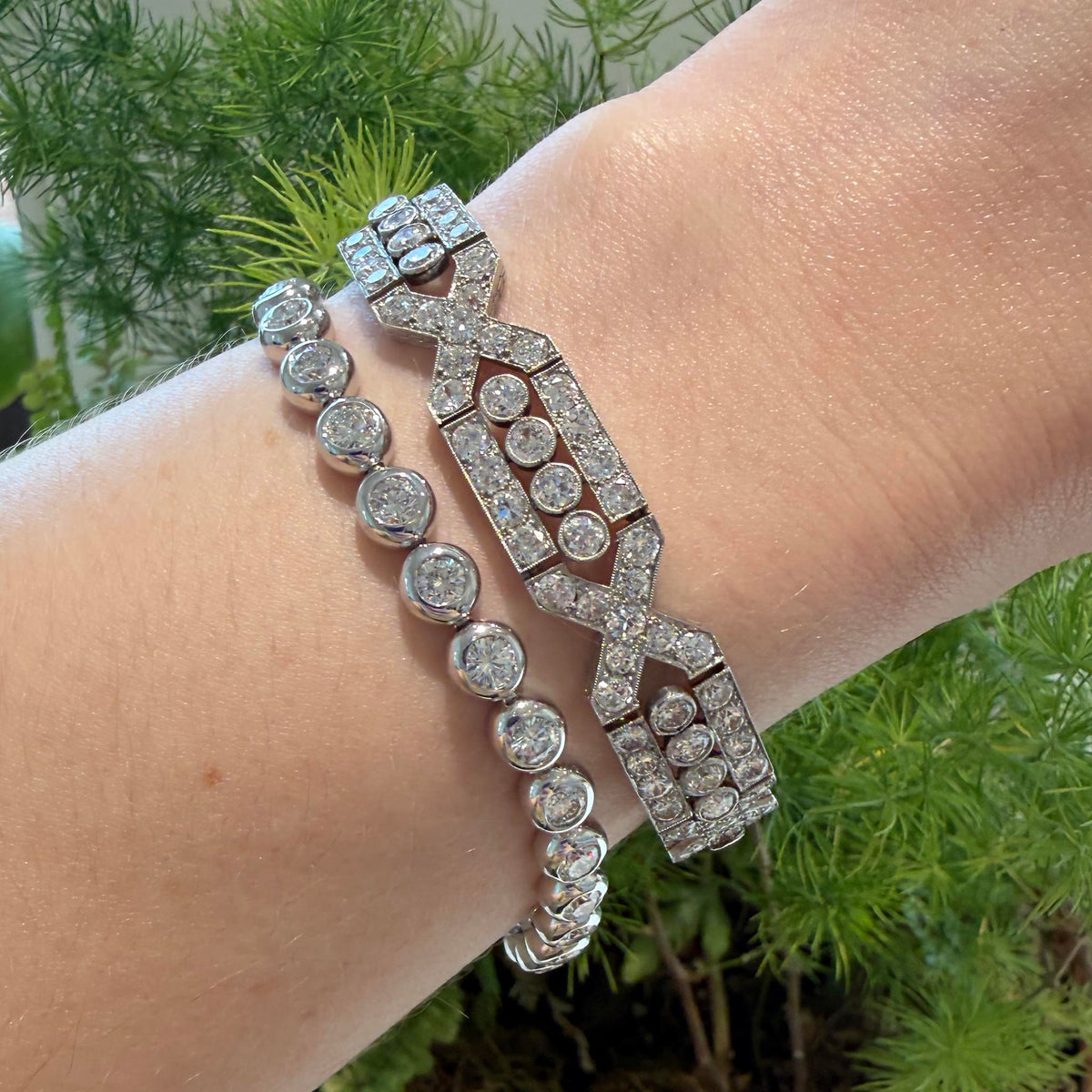 French Art Deco-style diamond and platinum bracelet worn on the wrist beside a bezel-set diamond bracelet. Estate bezel-set diamond tennis bracelet in 14K white gold worn on the wrist beside a wider Art Deco-style diamond bracelet.