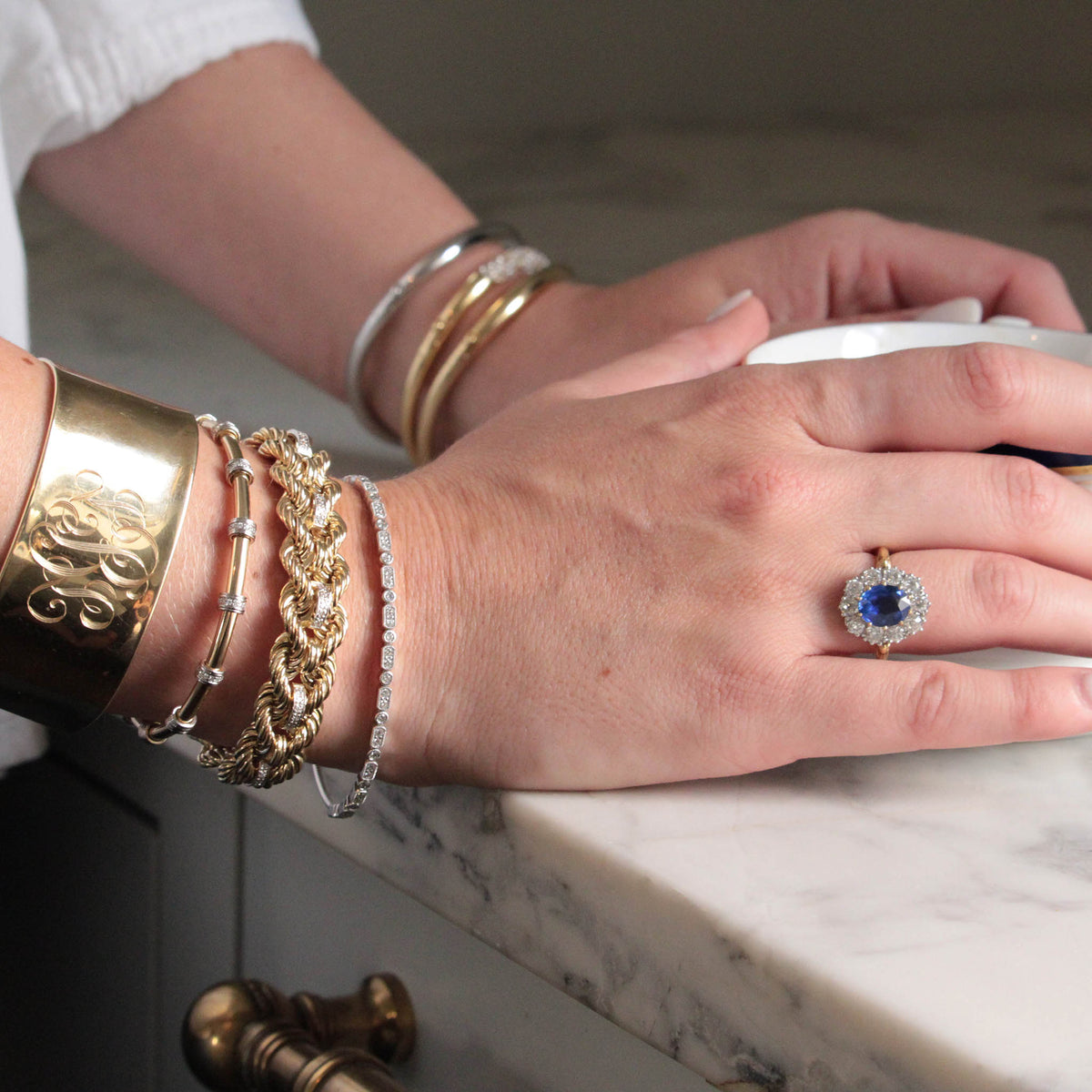 hands holding a coffee cup, wearing a Victorian-Style 2.56ct Oval Sapphire & Diamond Cluster Ring, stacked bangles and exclusive bracelets