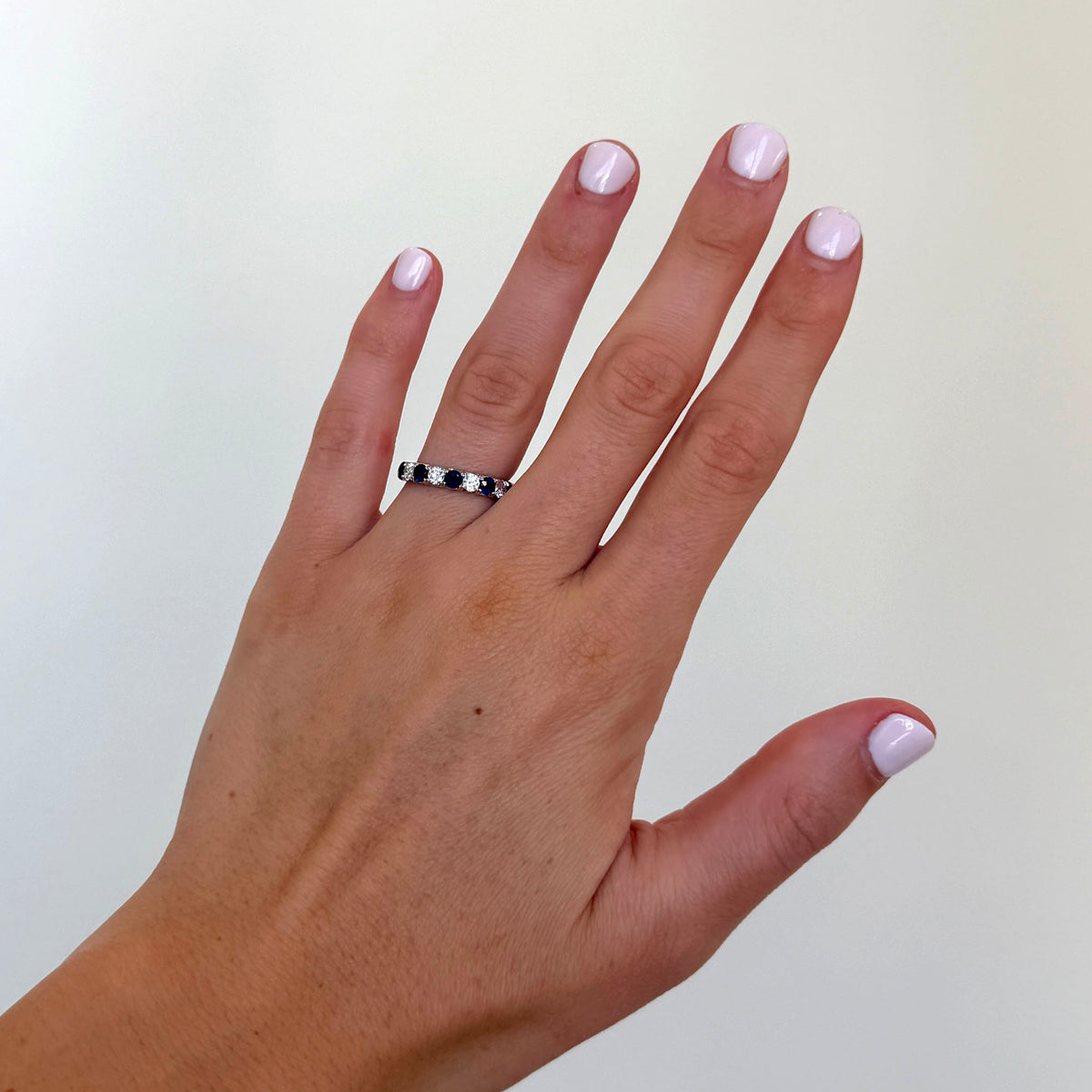A close-up of a hand wearing a band ring with small dark blue round stones, set against a plain off-white background.
