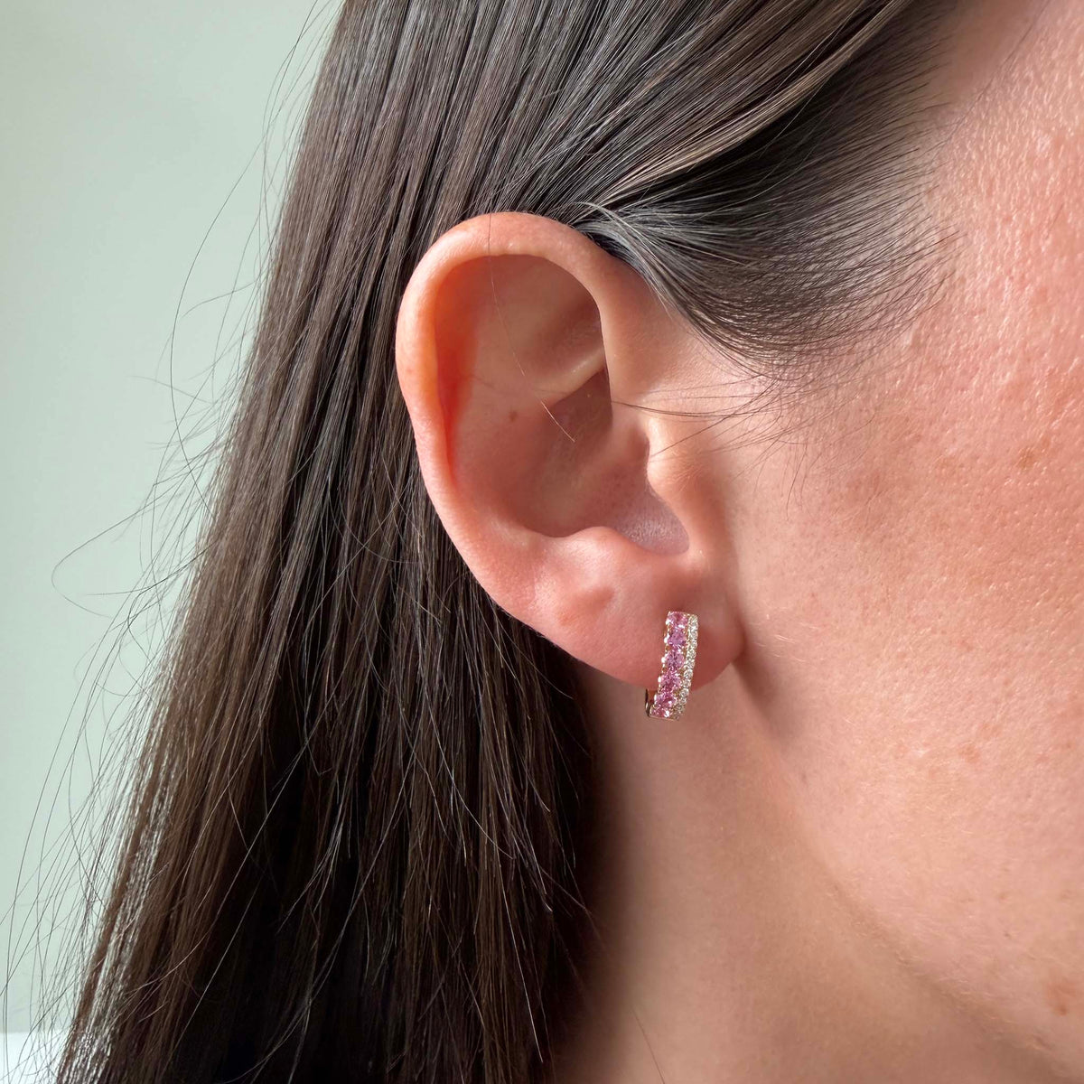 Pink sapphire and diamond huggie earring in 14K yellow gold worn close to the earlobe, showing its compact size and curved shape.