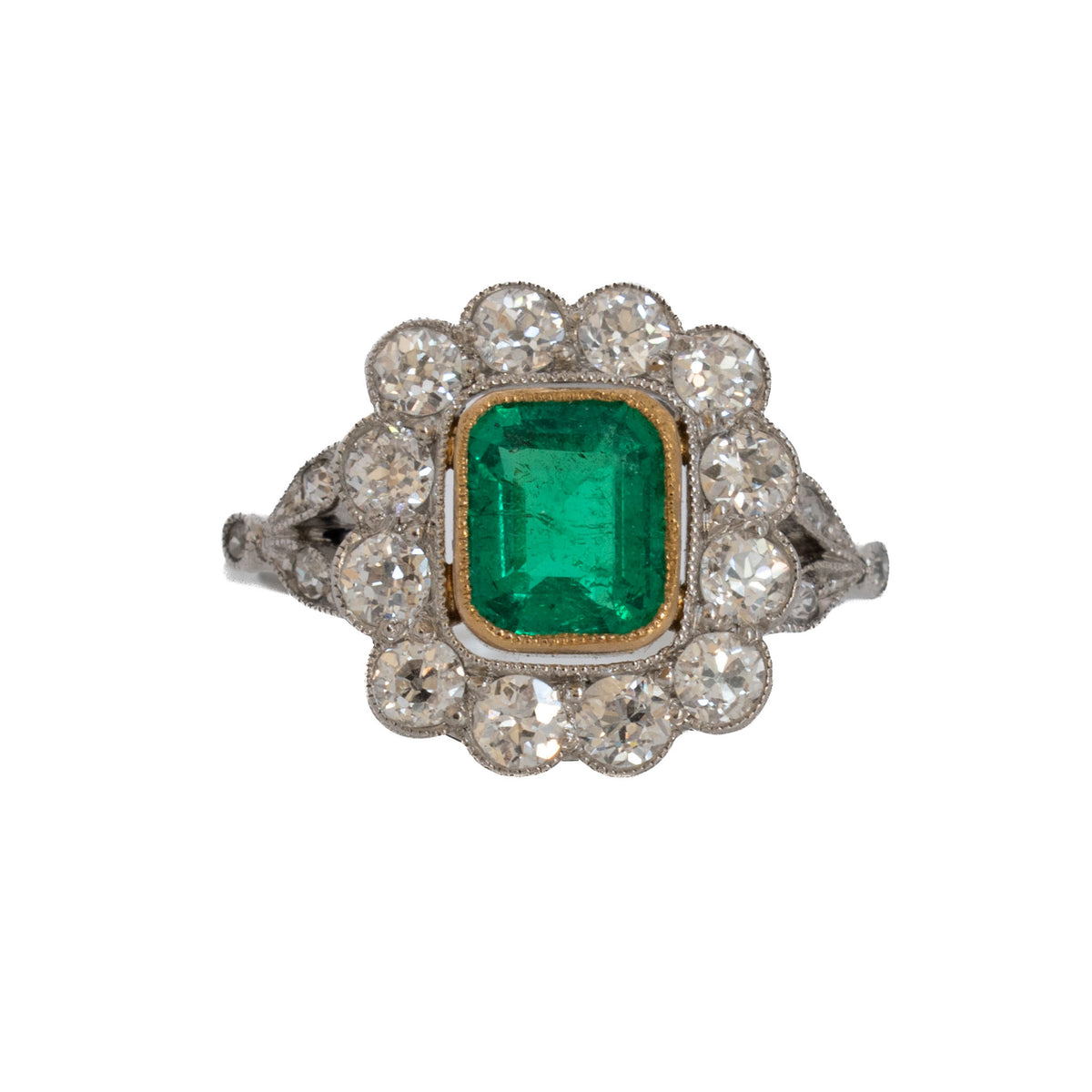 Edwardian emerald and diamond cluster ring with a rectangular green emerald in a yellow gold bezel, surrounded by round diamonds.