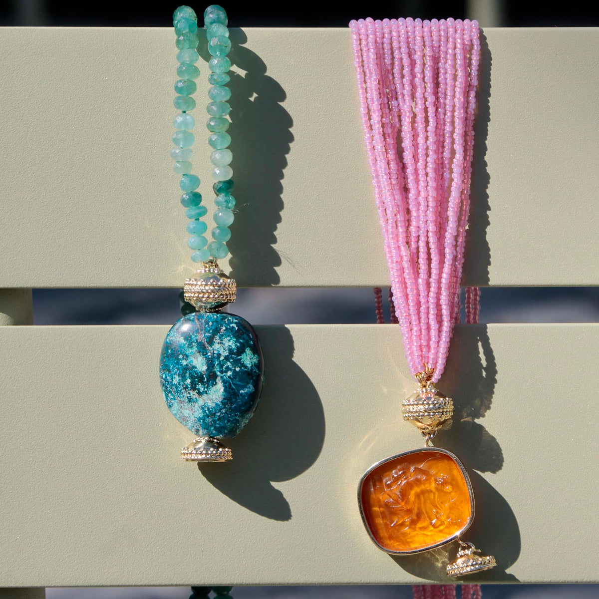 Clara Williams Large Green Chrysocolla Centerpiece on Grandidierite Rondelle Double-Strand Necklace next to Clara Williams Pink Ivory Coast Glass Multi-Strand Necklace with Diana Whisky Citrine Chiara Glass 14K Bezel Centerpiece draped over a light green chair