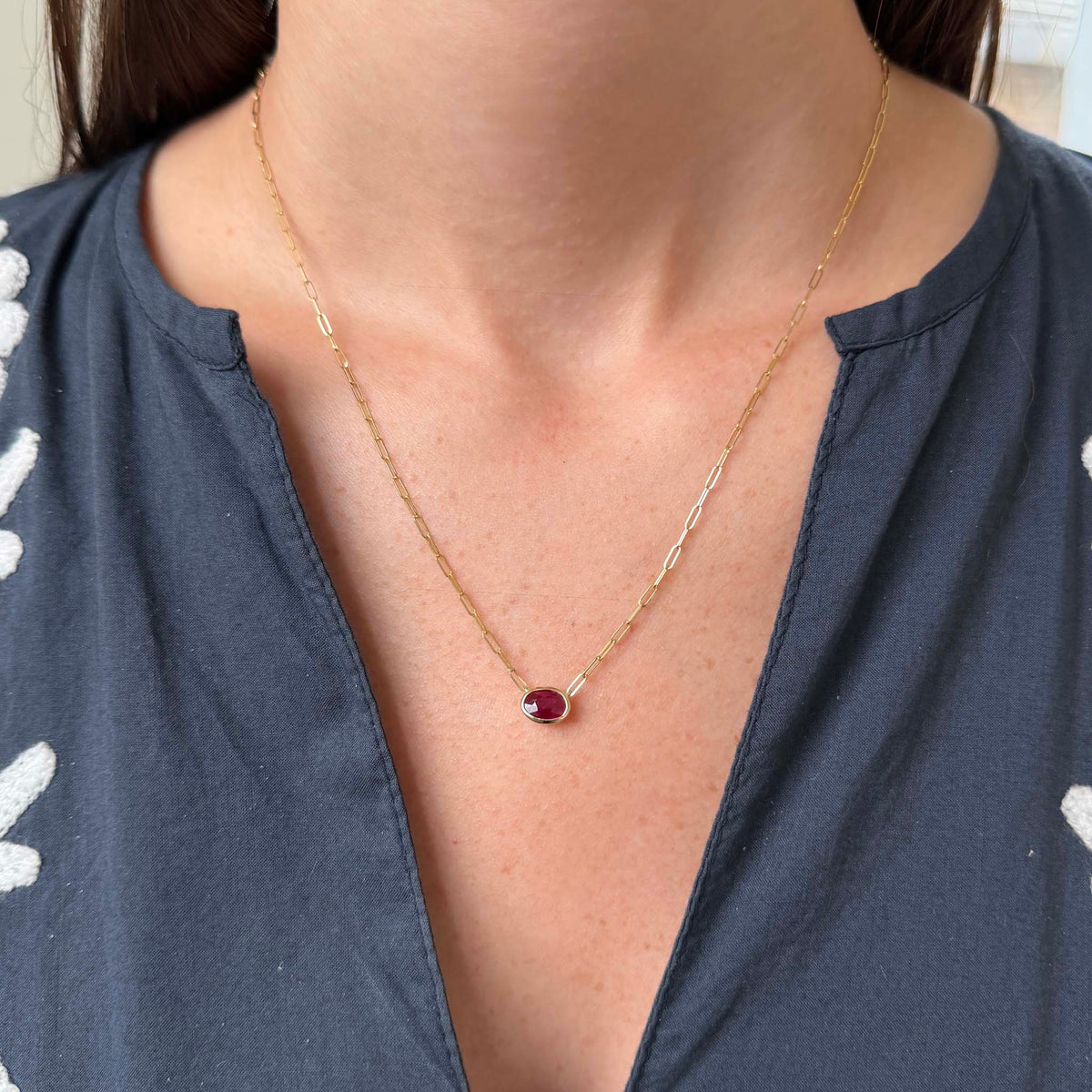 Oval ruby necklace in 14K yellow gold worn below the collarbone, showing the east-west bezel setting and delicate paperclip chain.