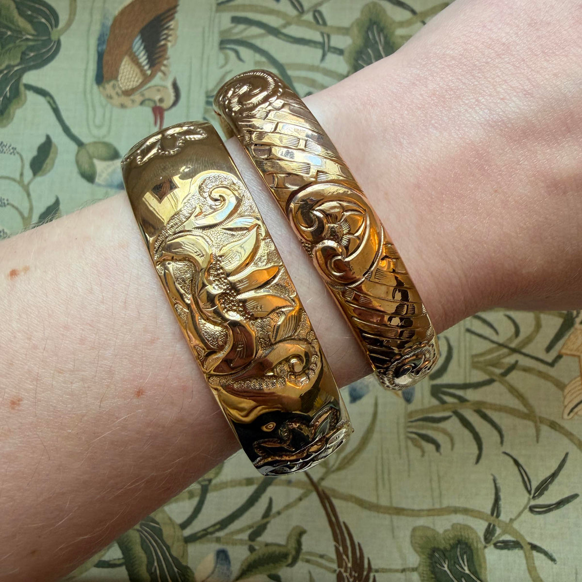 Model wrist wearing two Vintage Engraved Floral Pattern Gold-Filled Bangles over a pattern background.