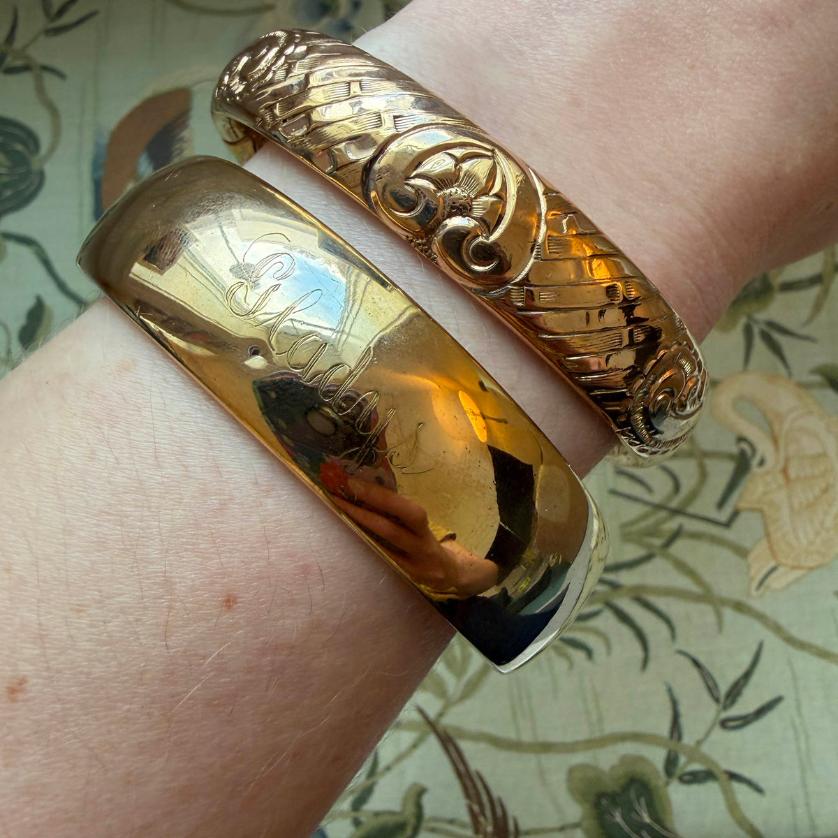 Model wrist wearing two Vintage Engraved Floral Pattern Gold-Filled Bangles, one bangle turned to the other side, over a pattern background.
