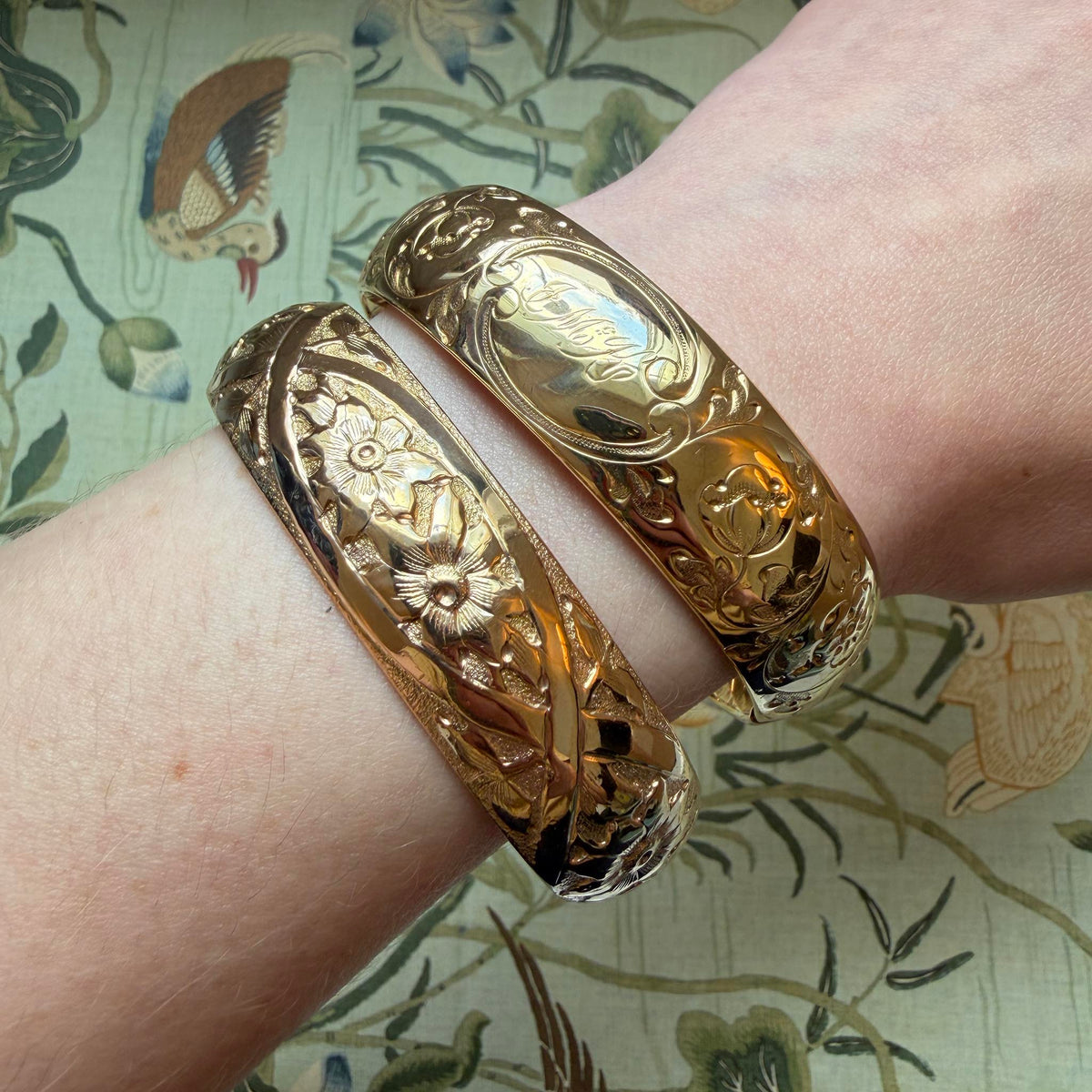 A model's wrist wearing two Vintage Engraved Floral Pattern Gold-Filled Bangles over a pattern background.