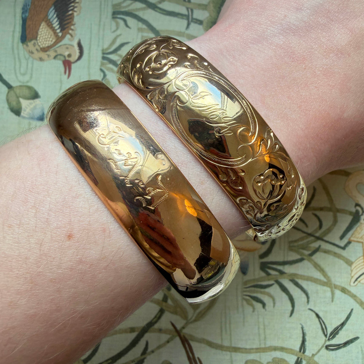 A model's wrist wearing two Vintage Engraved Floral Pattern Gold-Filled Bangles over a pattern background.