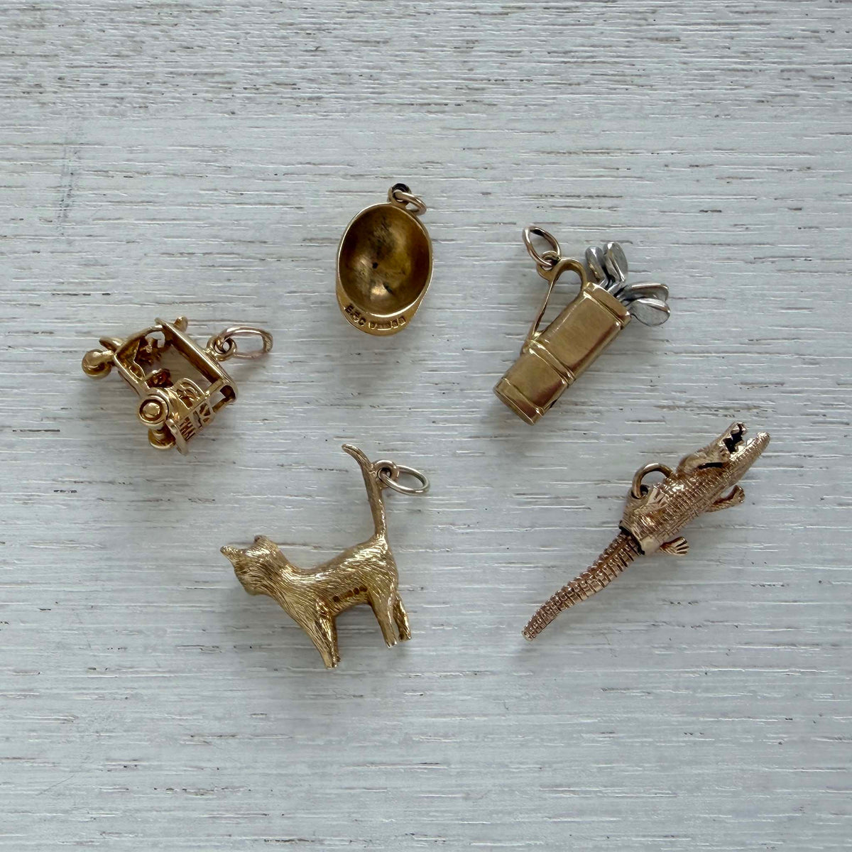 Underside of a vintage gold jockey cap charm beside golf cart, golf bag, cat, and crocodile charms on white wood.