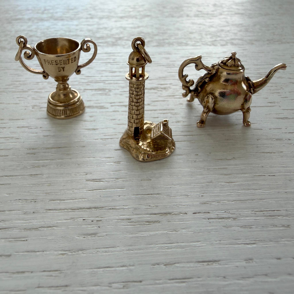 Vintage 9K yellow gold lighthouse and keeper’s house charm displayed between gold trophy and teapot charms on white wood.
