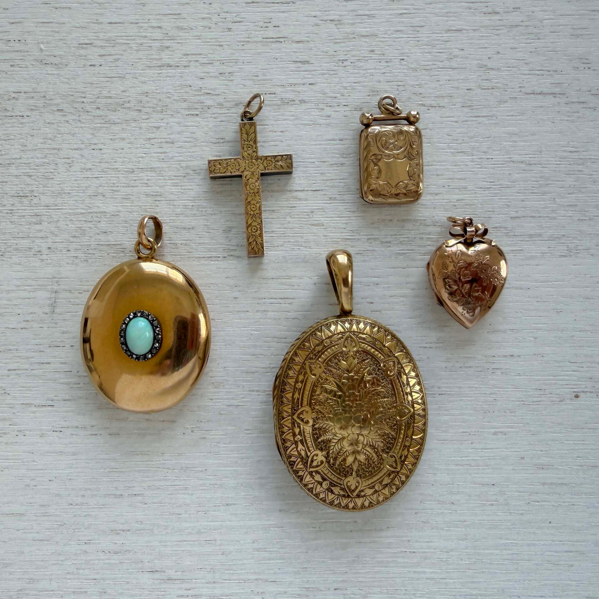 Edwardian engraved heart locket displayed with three other gold lockets and a cross pendant on white wood.
