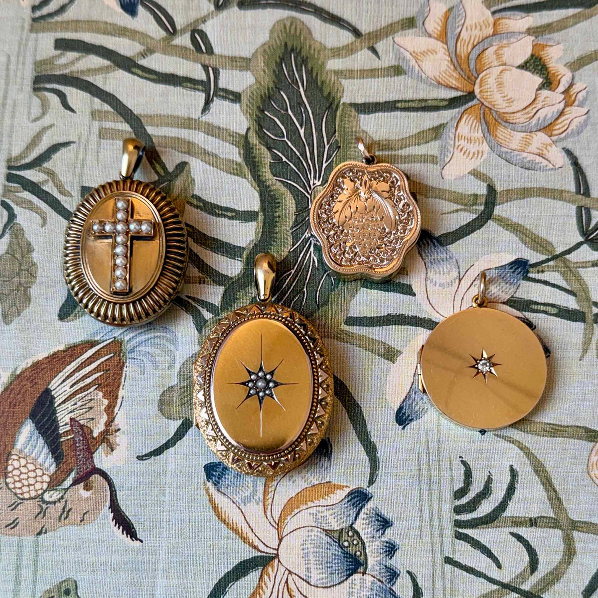 Four vintage gold-toned lockets and pendants displayed on a floral fabric background. The pendants feature ornate filigree borders, starburst details, a cross motif.