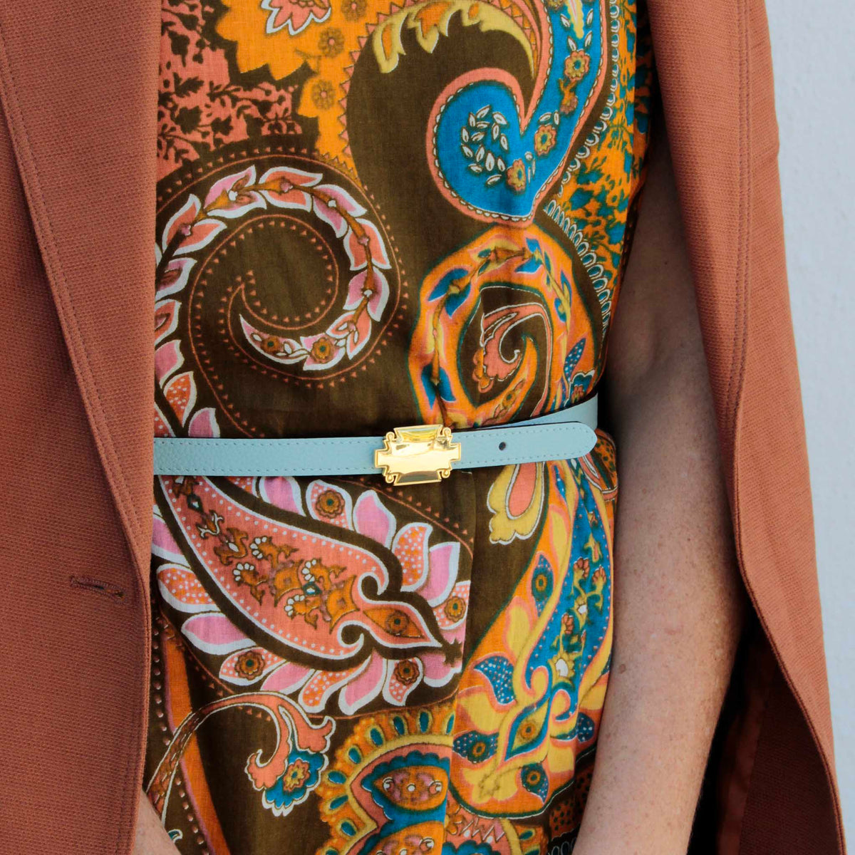 Light blue leather belt with a gold-plated plaque buckle, worn over a paisley print dress and a dusty rose blazer.