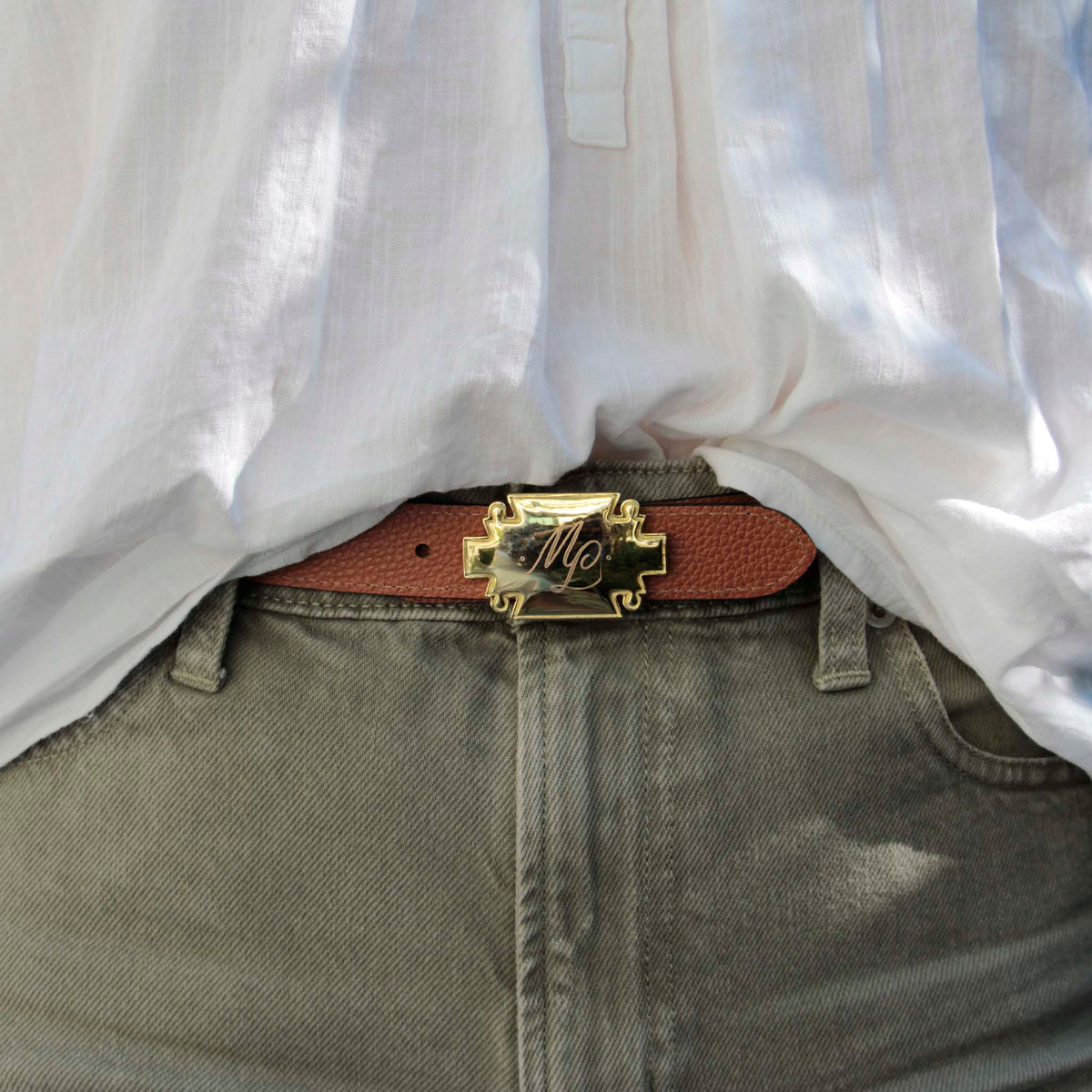 A close-up of a brown leather belt with a gold-plated decorative buckle featuring engraved initials ML, worn with green jeans and a white shirt.
