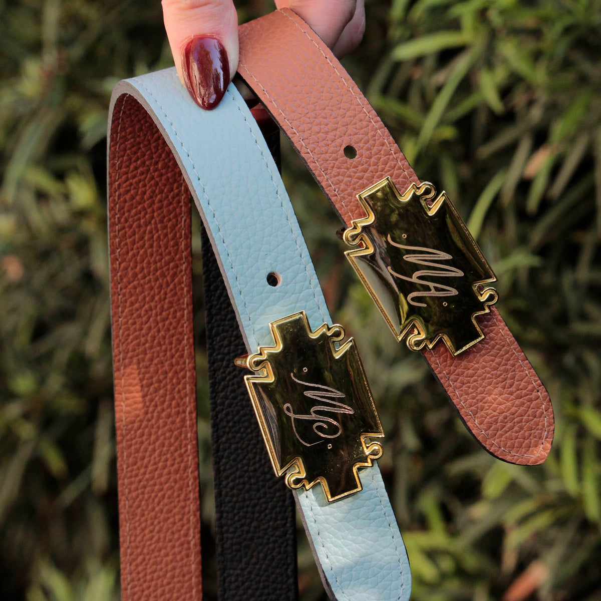 Hand holding two belts in saddle brown, and light blue with gold monogrammed plaque buckles against greenery.